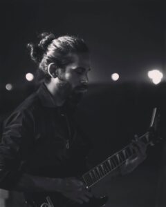 A black-and-white, artistic medium shot of a male guitarist with his hair tied back, deeply focused while playing an electric guitar outdoors at night against a blurred background of city lights.