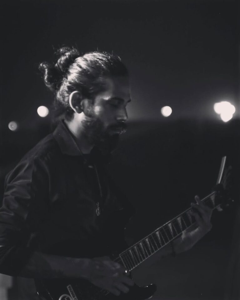 A black-and-white, artistic medium shot of a male guitarist with his hair tied back, deeply focused while playing an electric guitar outdoors at night against a blurred background of city lights.